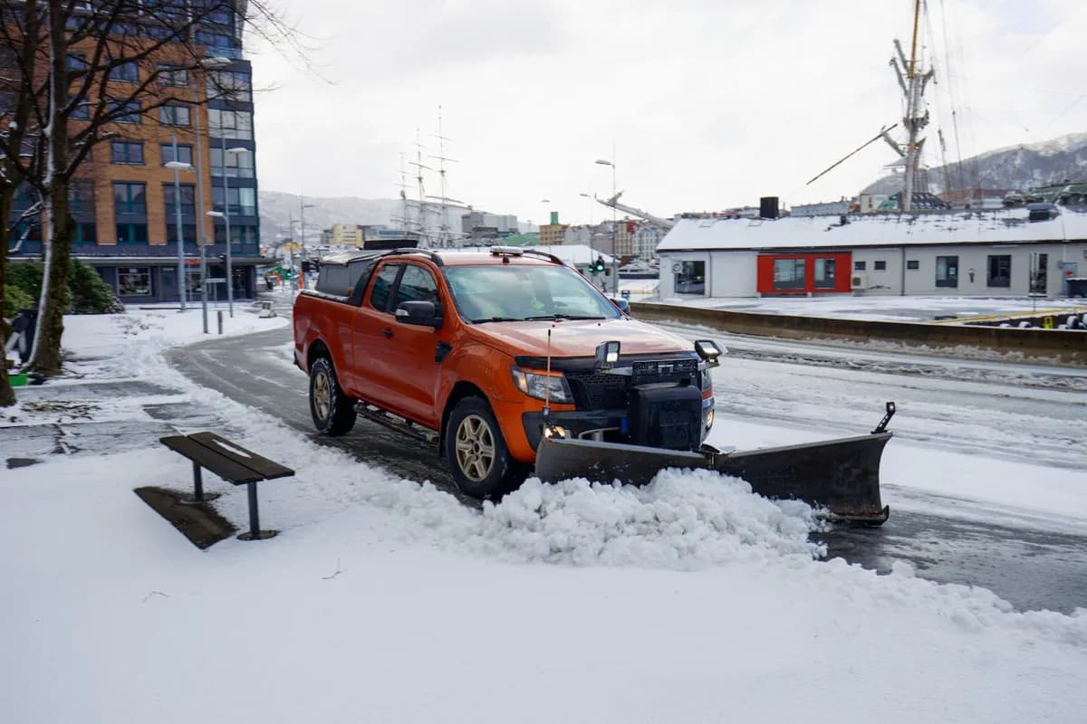 Orangefärgad pickup med snöplog röjer snö på en gata i en stadsmiljö.