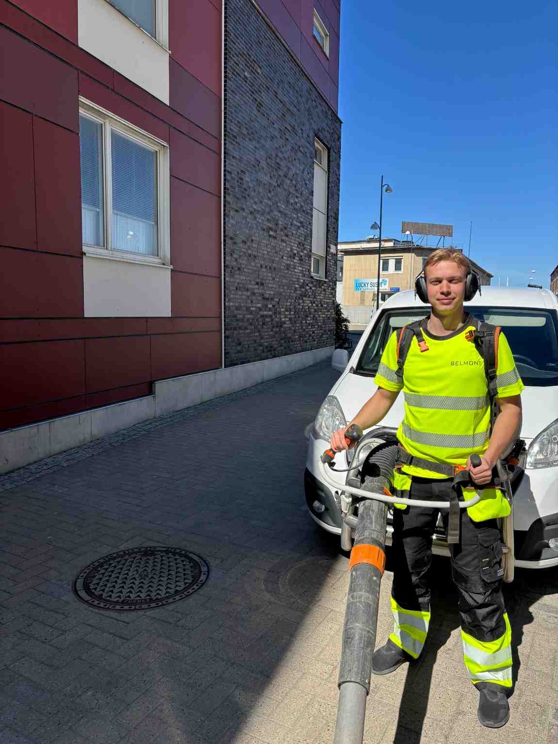 Medarbetare från Belmont i varselkläder står med arbetsutrustning bredvid ett tjänstefordon på en gata.